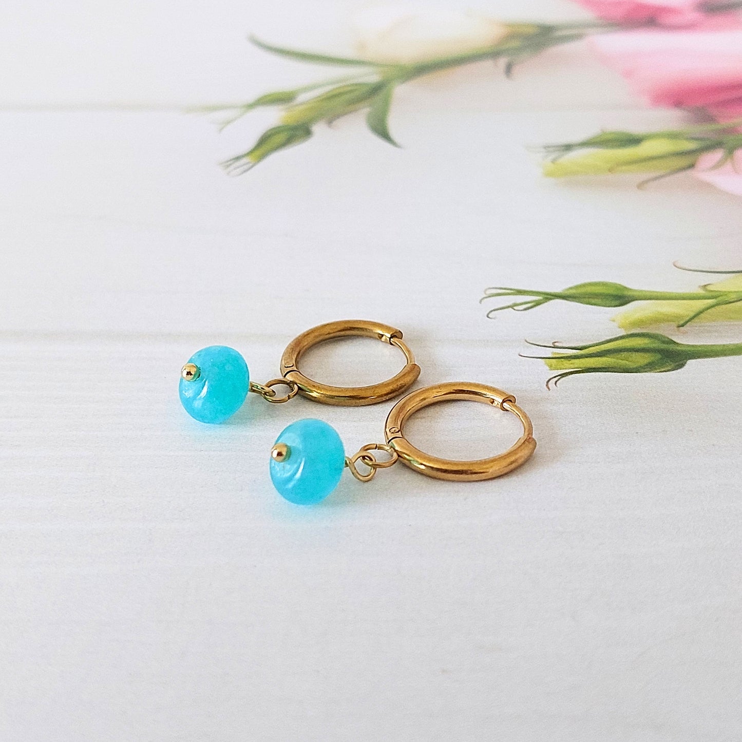 A pair of gold-plated stainless steel hoop earrings with small, rondelle shaped blue turquoise jade bead drops, displayed on a white wooden surface with soft pink flowers in the background.