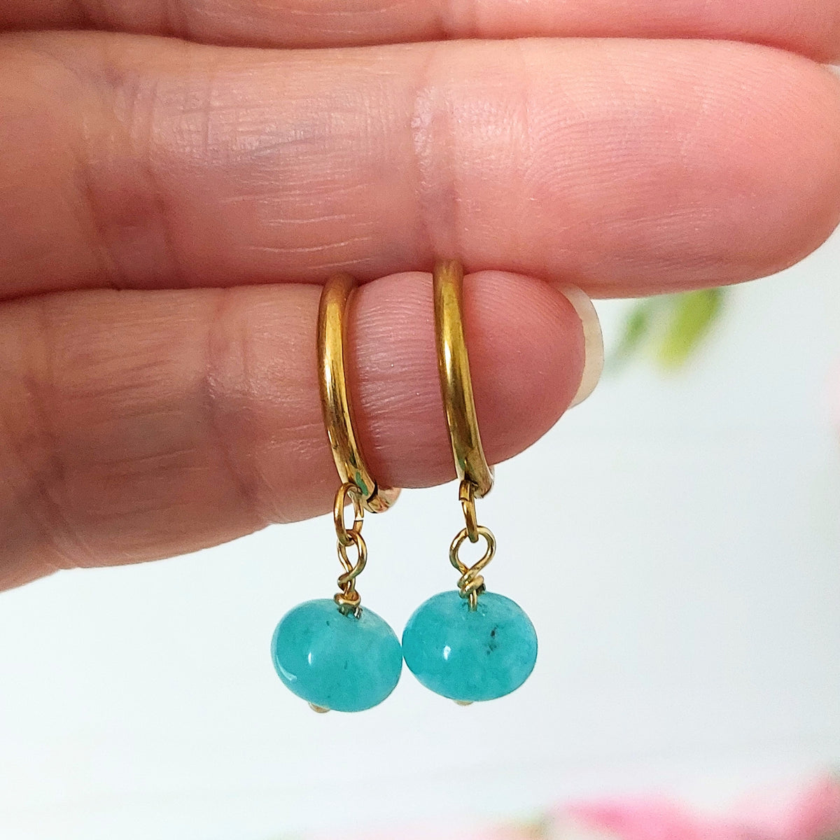 A pair of gold-plated stainless steel hoop earrings with oval turquoise-blue jade gemstone drops, held between a person's fingers against a soft white background.