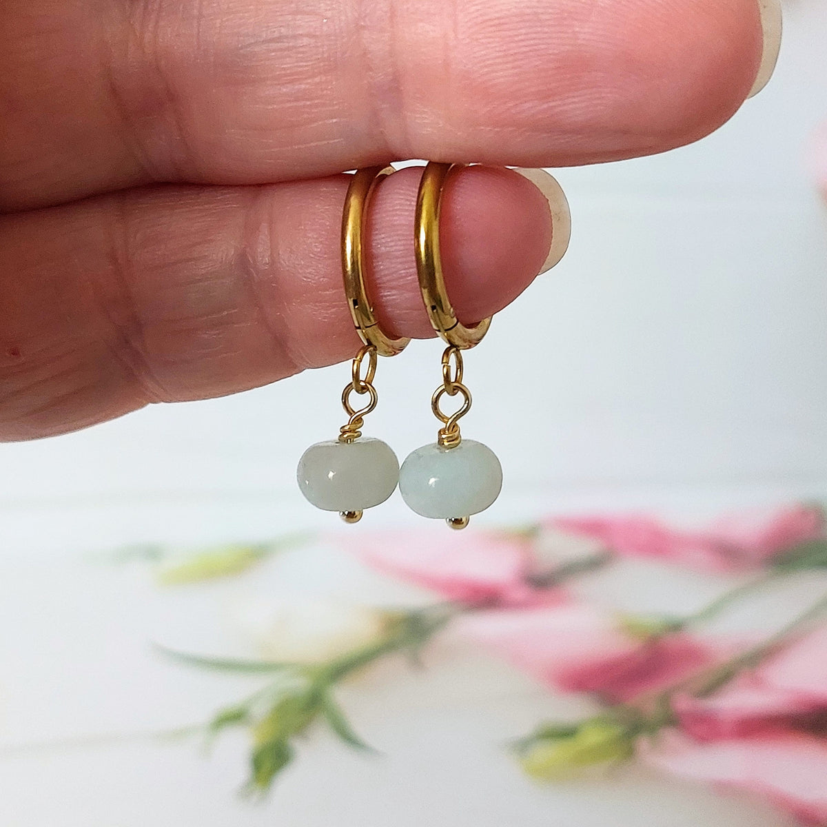 A close-up of a hand holding a pair of gold-plated stainless steel hoop earrings with dangling light blue flower amazonite beads, against a soft-focus background with pink flowers.