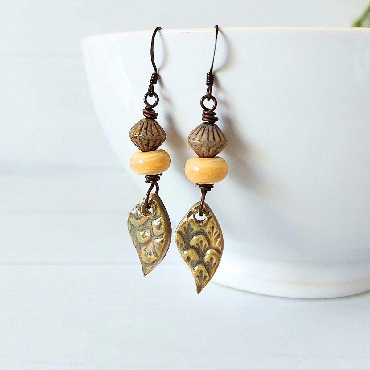 A pair of bohemian-style dangle earrings with olive ceramic decorative charms, light yellow lampwork glass beads, and dark olive textured beads, hanging from a white ceramic bowl on a white wooden surface.