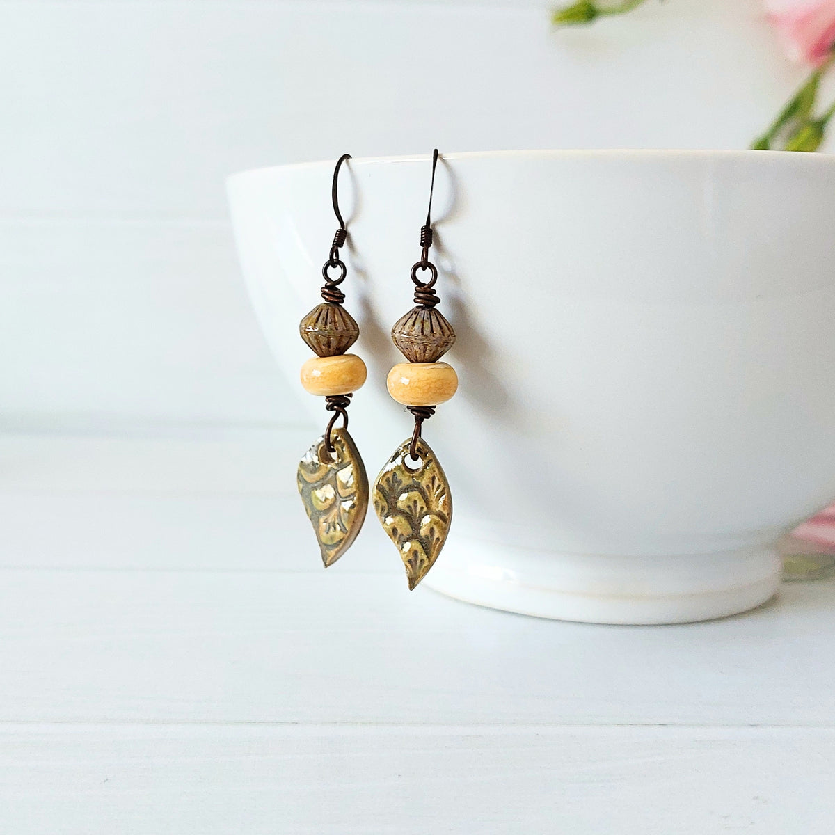 A pair of bohemian-style dangle earrings with olive ceramic decorative charms, light yellow lampwork glass beads, and dark olive textured beads, hanging from a white ceramic bowl on a white wooden surface.