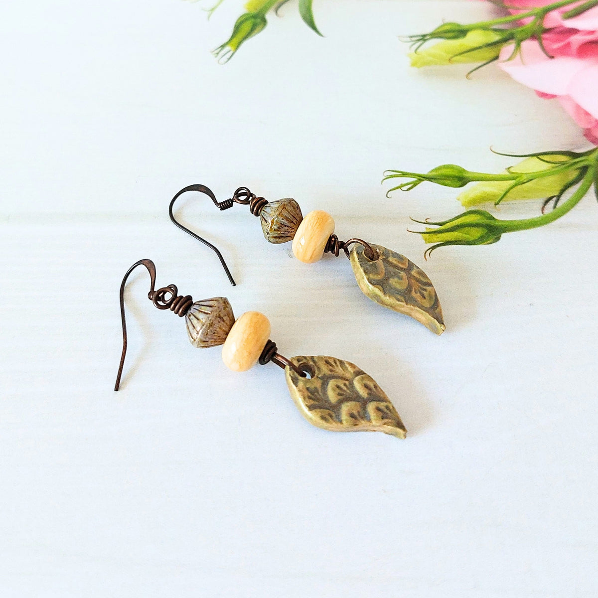 A pair of artisan drop earrings with olive ceramic tear-drop charms, light yellow lampwork glass beads, and dark olive bicone beads, on a white wooden surface.