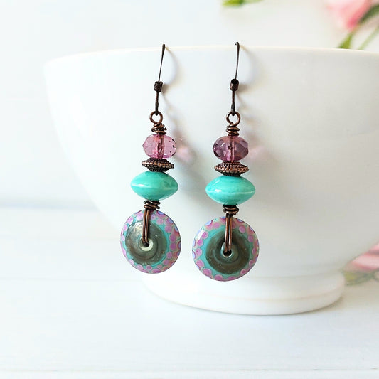A pair of handmade dangle earrings with mint green and purple beads, displayed on a white ceramic bowl against a light background.