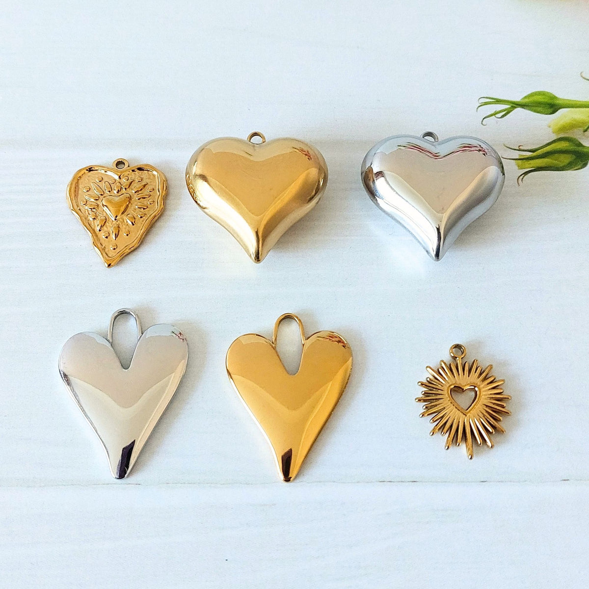 Six heart-shaped metal jewelry charms in polished gold and silver finishes, arranged on a white wooden surface with a small green flower stem in the corner.