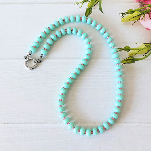 A beaded necklace with light mint green oval beads separated by tiny pink beads, featuring a silver-toned clasp, displayed on a white wooden surface with decorative flowers in the corner.