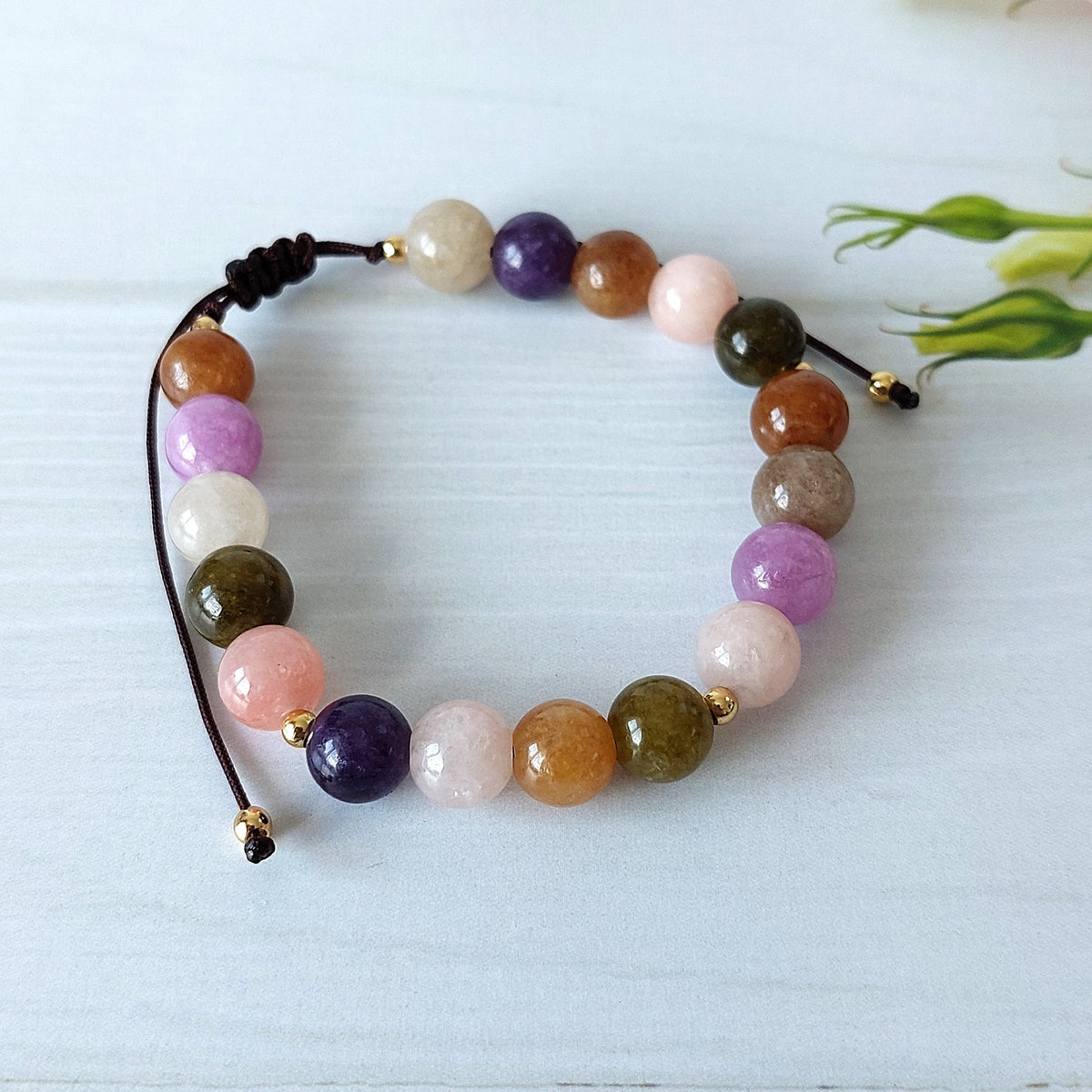 A multicolor beaded bracelet made of polished round gemstone beads in shades of purple, pink, orange, green, brown, and white, strung on a dark cord with small gold spacer beads, displayed on a light blue wooden surface.