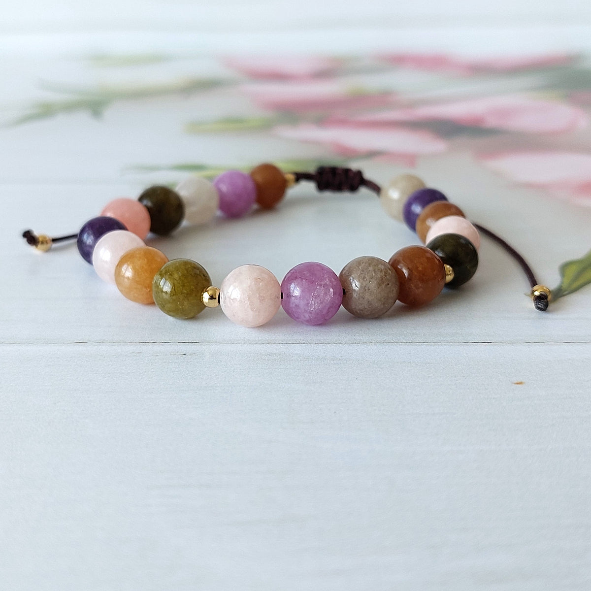 A beaded cord bracelet with polished multicolor gemstone beads in shades of purple, pink, brown, green, and cream, strung on a dark brown cord with gold-toned metal tips, displayed on a light wooden surface.