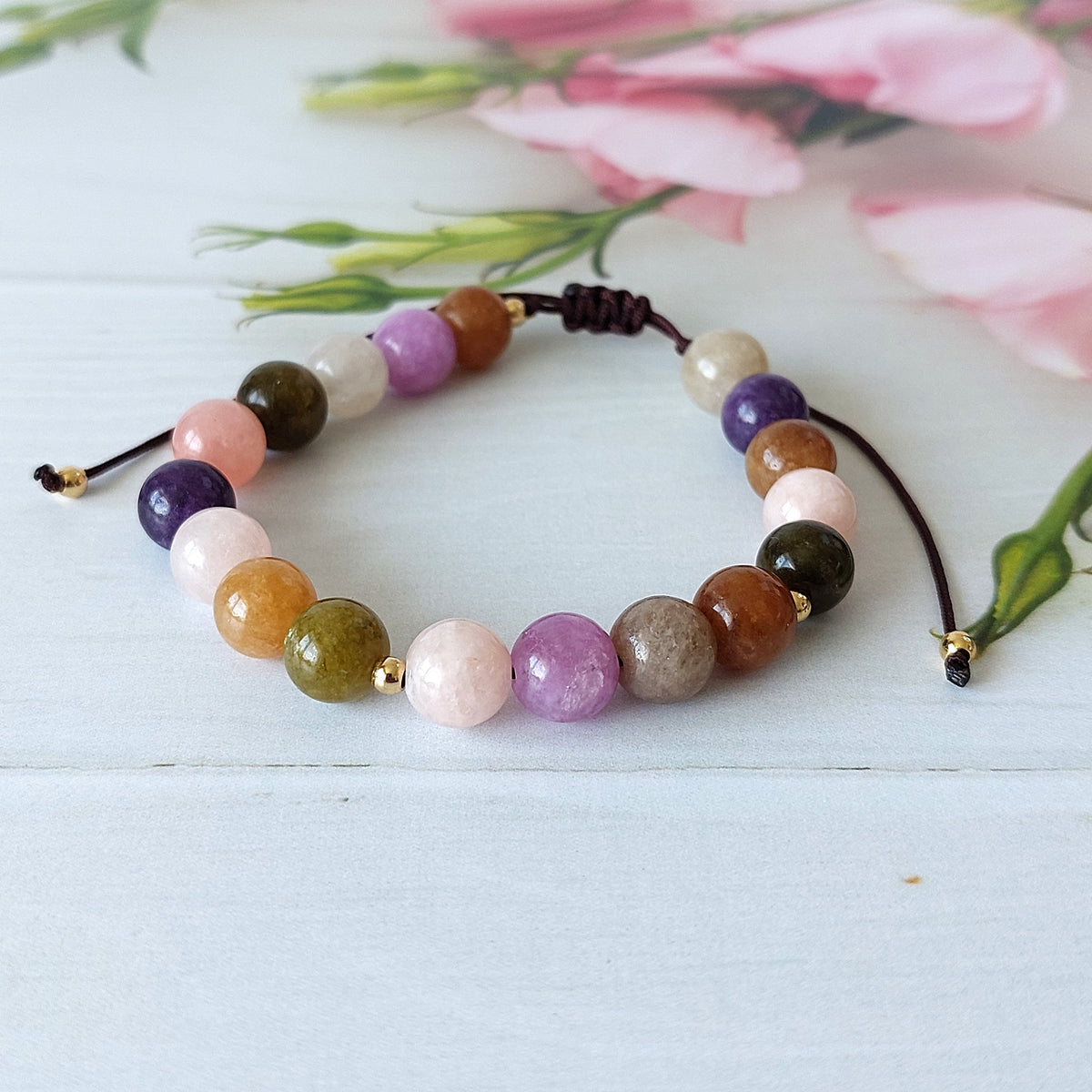 A beaded jade bracelet made of polished round gemstone beads in multiple colours including pink, purple, brown, green, and white, strung on a dark cord with gold spacer beads, displayed on a light wooden surface with soft pink flowers in the background.