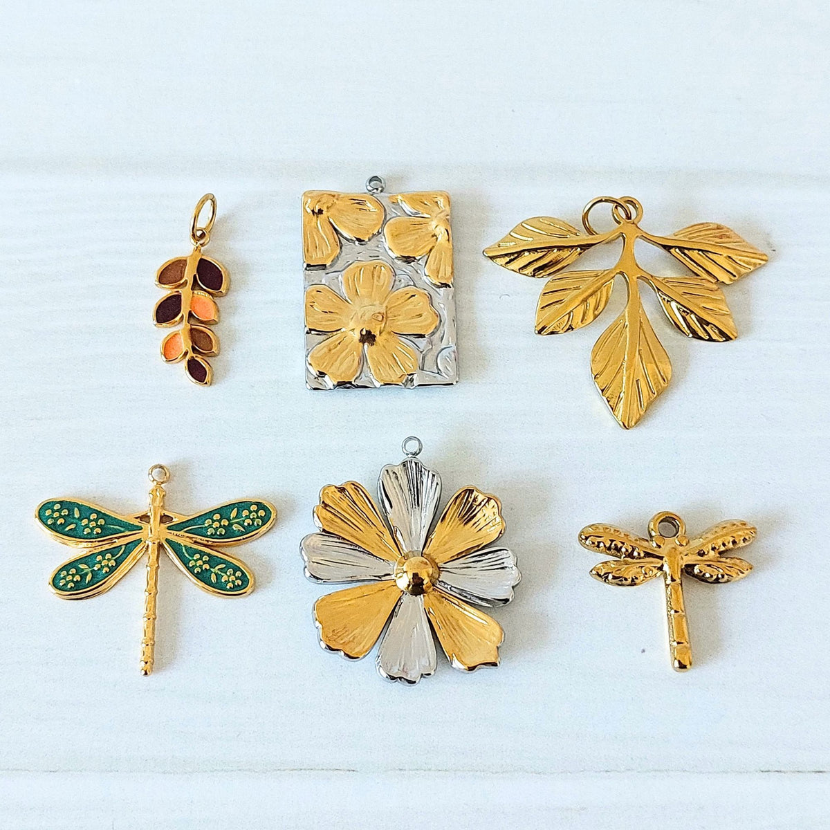 Six jewellery pendants arranged on a white textured background: a gold-tone enamel leaf pendant, a gold & silver-tone floral rectangular pendant, a gold-tone leaf branch pendant, a turquoise-and-gold dragonfly pendant, a gold-and-silver-tone flower pendant, and a gold-tone dragonfly pendant.
