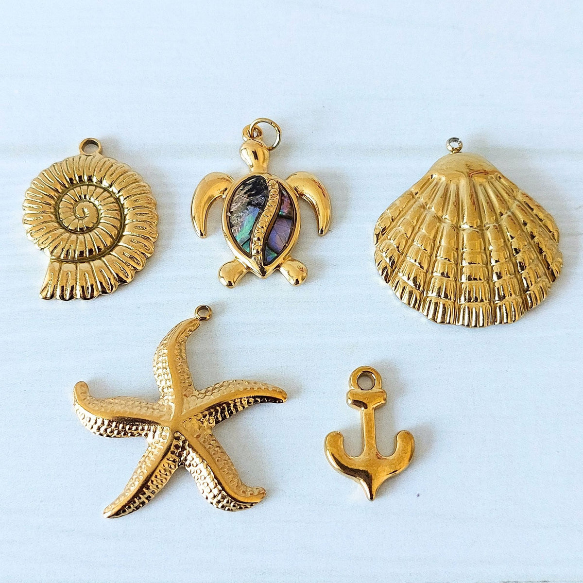 Five gold-toned nautical-themed jewelry charms on a light blue background: a spiral shell, a sea turtle with an abalone shell, a scallop shell, a starfish, and an anchor.