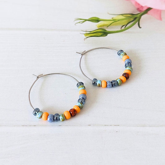 A pair of silver-tone stainless steel hoop earrings with small multicoloured beads in orange, blue, and grey, displayed on a white wooden surface with a soft pink flower bud in the background.