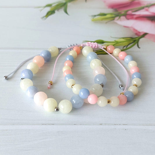 Two pastel beaded bracelets with pink cords, featuring round jade beads in light blue, pale pink, cream, and white, accented with small gold spacers, arranged on a white wooden surface with pink flowers in the background.
