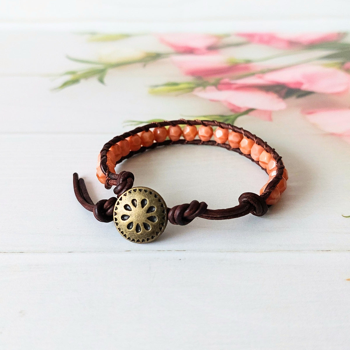 A handmade beaded wrap bracelet with dark brown leather cords, soft coral beads, and a decorative brass metal button fastener, displayed on a white wooden surface with a soft pink floral background.