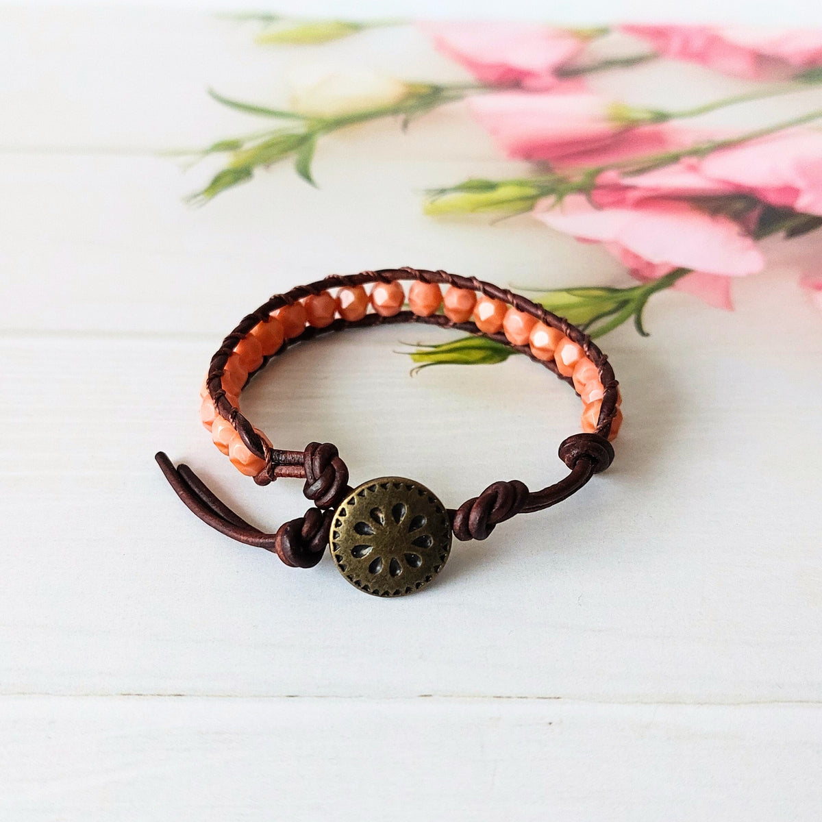 A handmade beaded wrap bracelet with dark brown leather cords, soft coral beads, and a decorative brass metal button fastener, displayed on a white wooden surface with a soft pink floral background.