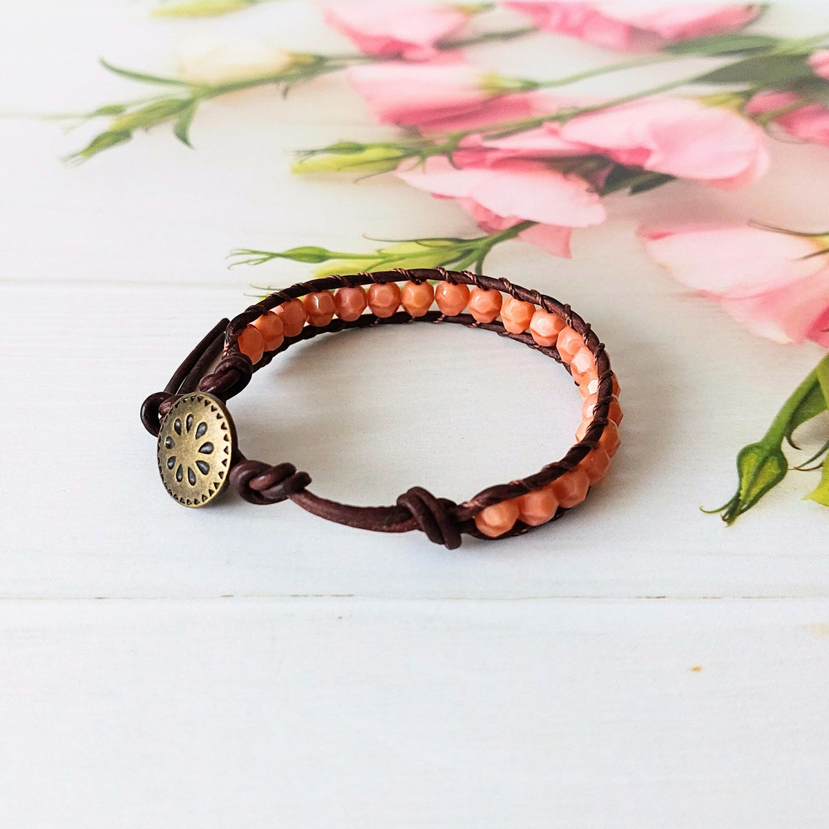 A handmade leather bracelet with small peach-coloured beads and a round antique brass metal button clasp, displayed on a white wooden surface with pink flowers in the background.