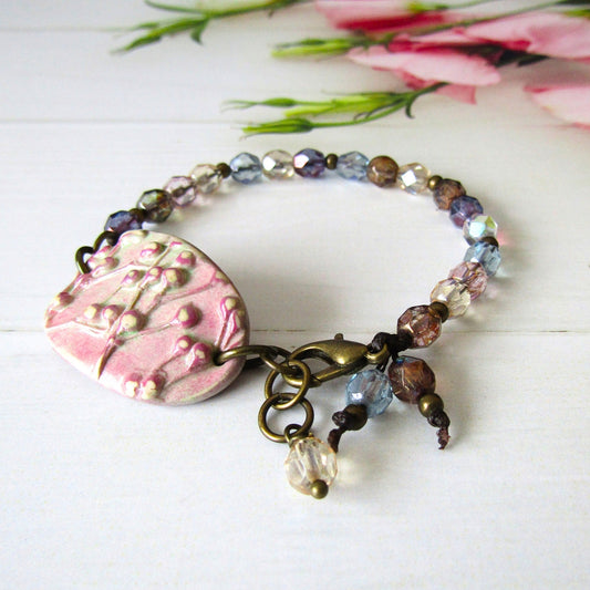 A handmade beaded bracelet with a pink textured polymer clay connector charm, antique brass hardware, and faceted beads in pink, blue, and brown, displayed on a white wooden surface with blurred pink flowers in the background.