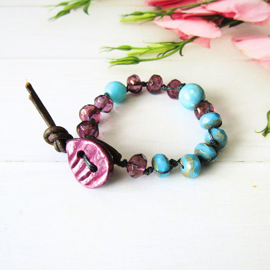 A handmade beaded bracelet featuring turquoise and purple beads, a handmade ceramic purple petal shaped button, and a brown leather toggle clasp, displayed on a white surface with blurred pink flowers in the background.