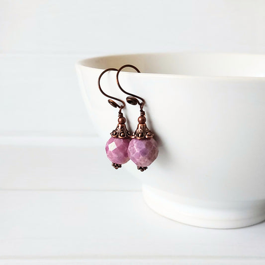 A pair of dangling earrings with faceted light purple beads and dark copper hooks, resting against a white ceramic bowl on a white surface.