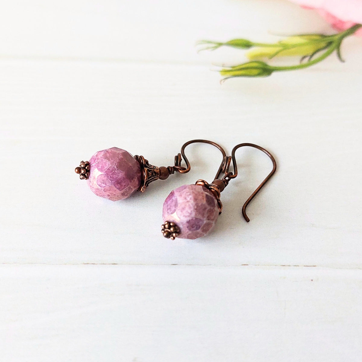 A pair of handmade dangle earrings with faceted light purple beads and copper-colored wire hooks, displayed on a white wooden surface with a soft pink flower in the background.