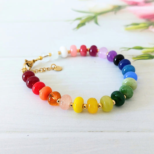 A gemstone beaded bracelet arranged in a rainbow sequence with gold-tone stainless steel spacer beads and a gold clasp, photographed on a light wood surface with soft blurred flowers in the background.