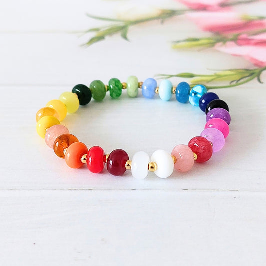 A beaded stretch bracelet arranged in a rainbow sequence of colorful round beads with small gold-tone stainless steel spacer beads, displayed on a white wooden surface with soft pink flowers blurred in the background.