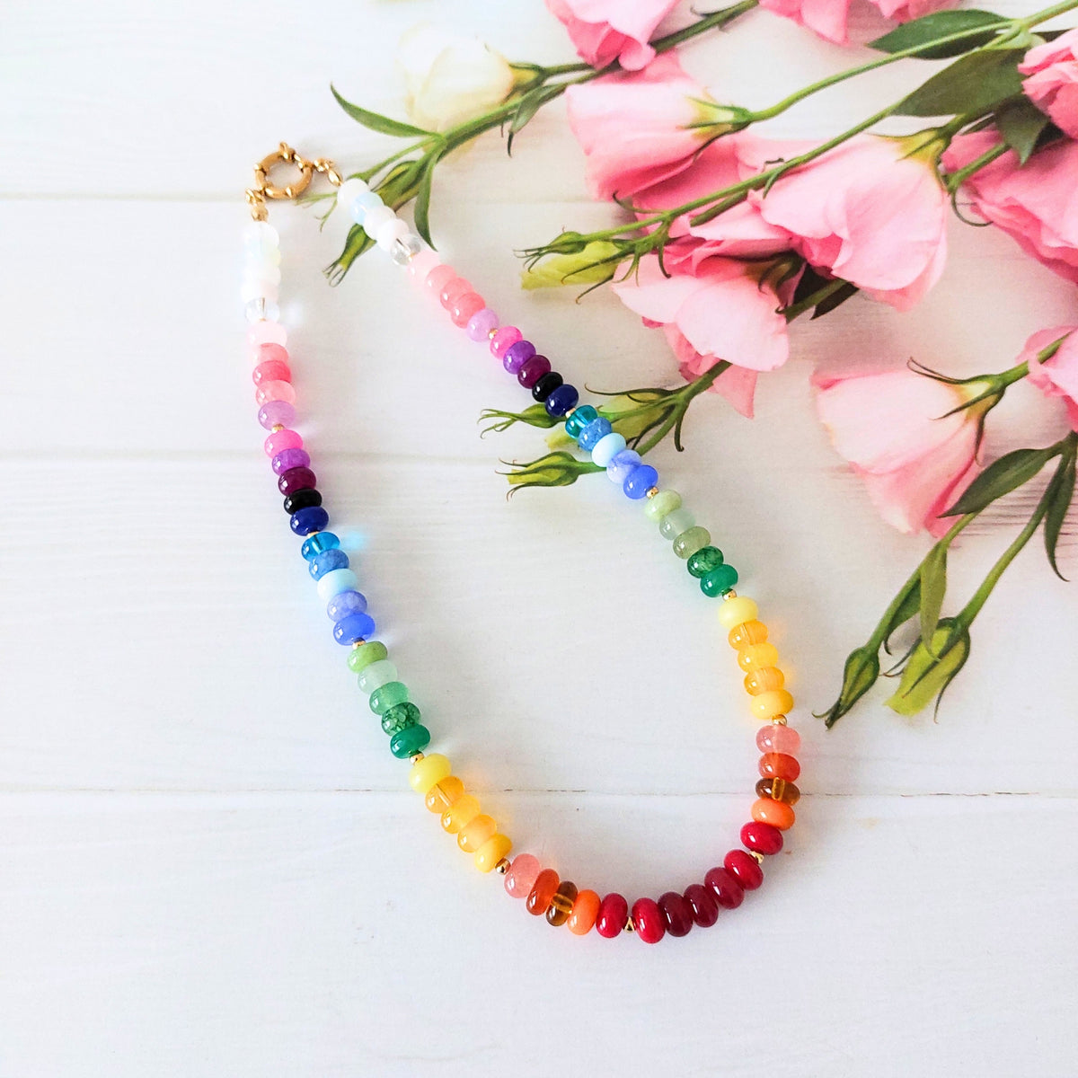 A beaded necklace arranged in an oval shape on a white wooden surface, decorated with pink flowers in the upper right corner. The necklace features a gradient of colorful rondelle beads transitioning through rainbow shades, with a gold clasp at the top.