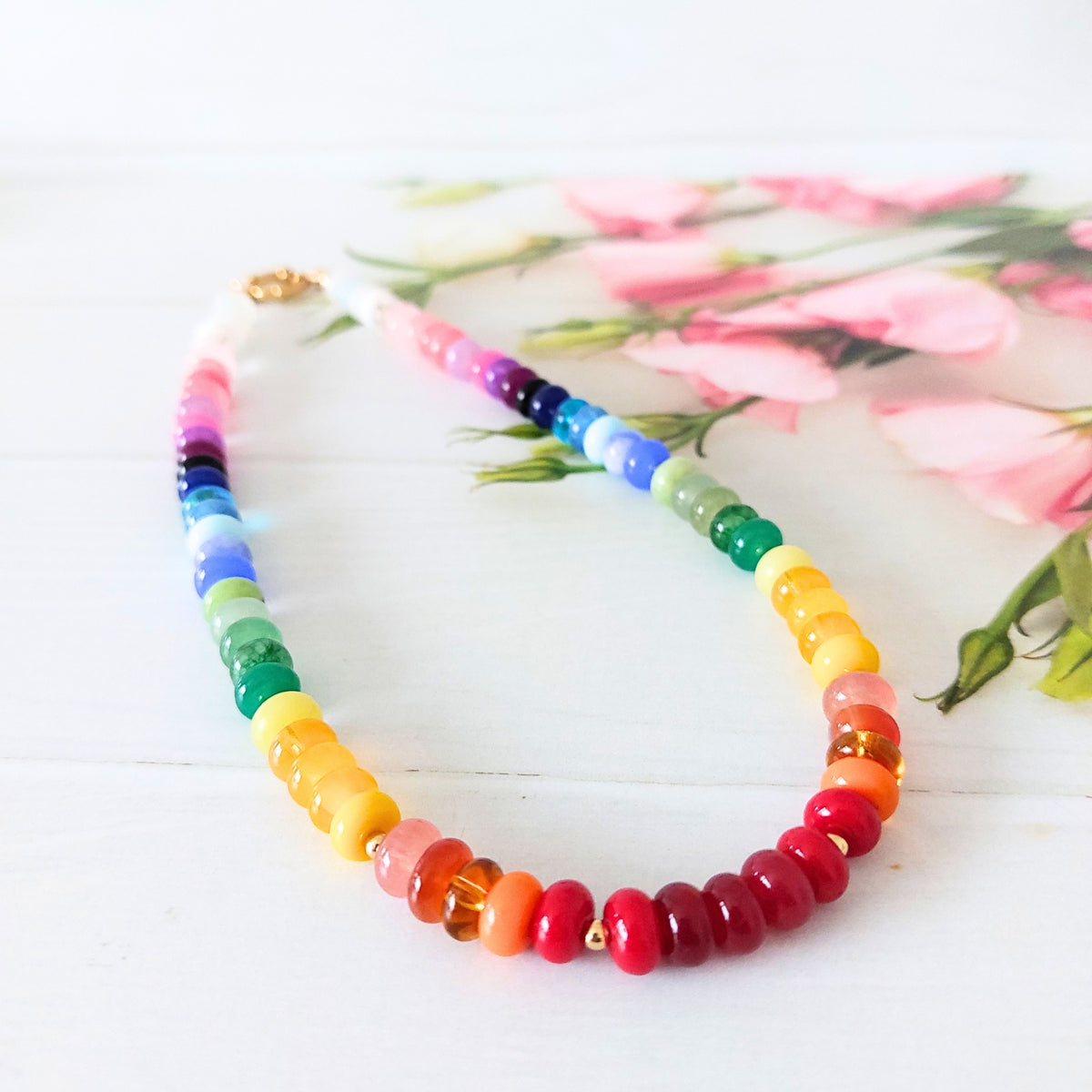 A beaded necklace with a rainbow colour sequence, featuring translucent round beads in shades of pink, purple, blue, green, yellow, orange, and red, with a gold clasp, displayed on a white wooden surface with pink flowers in the background.