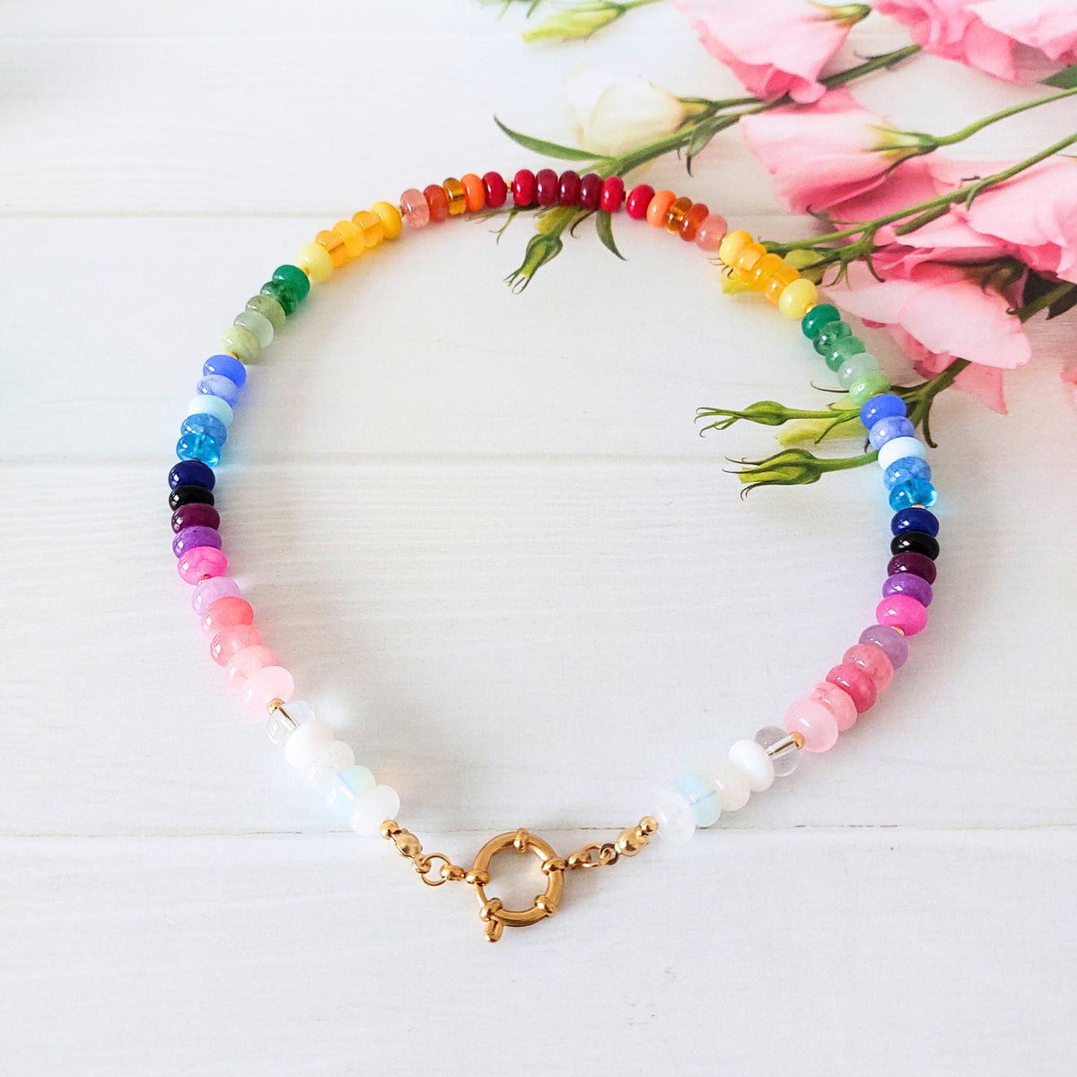 A beaded necklace with a rainbow of translucent gemstone beads, featuring a gold clasp, displayed on a white wooden surface next to pink flowers.