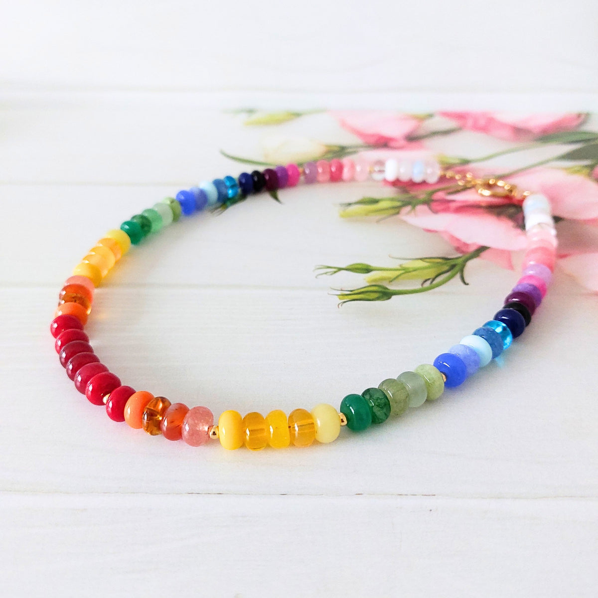 A beaded rainbow necklace arranged in a circle on a white wooden surface with pink flowers in the background. The beads transition through red, orange, yellow, green, blue, purple, and pink, with a staement gold clasp visible at the top.