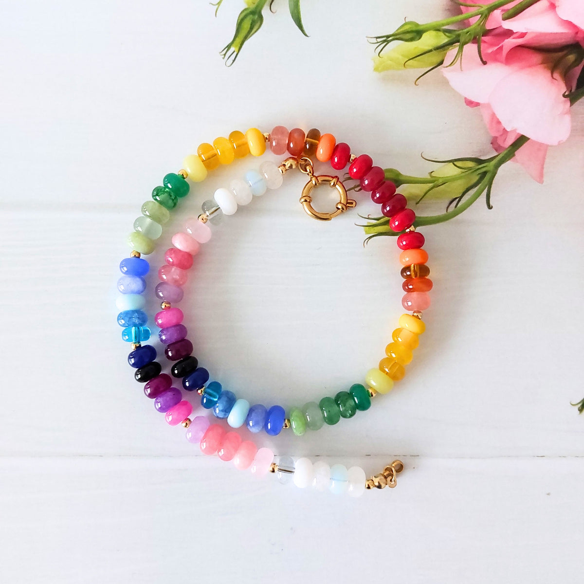 A rainbow beaded necklace with gold-toned accent beads, arranged on a white wooden surface next to pink flowers.