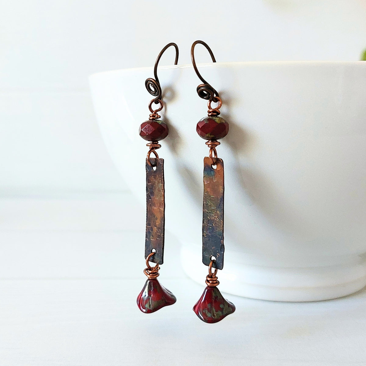 A pair of handmade dangle earrings with dark copper hooks, a faceted burgundy bead, a rectangular copper plate showing brown and blue patina, and a teardrop-shaped burgundy bead at the bottom, displayed on a white ceramic bowl.