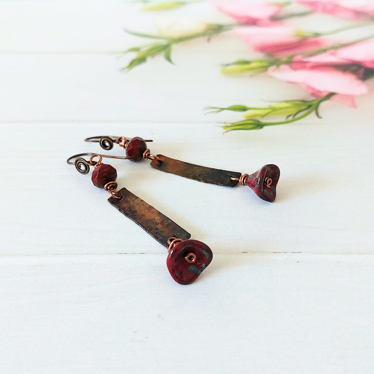 A pair of handmade dangle earrings with dark brown rectangular copper bars, copper wire wrapping, and deep red marbled beads, displayed on a white wooden surface with soft pink flowers in the background.