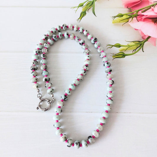 A beaded necklace with grey beads featuring black and pale blue marbled patterns and bright pink accents, displayed on a white wooden surface with pink flowers in the corner.