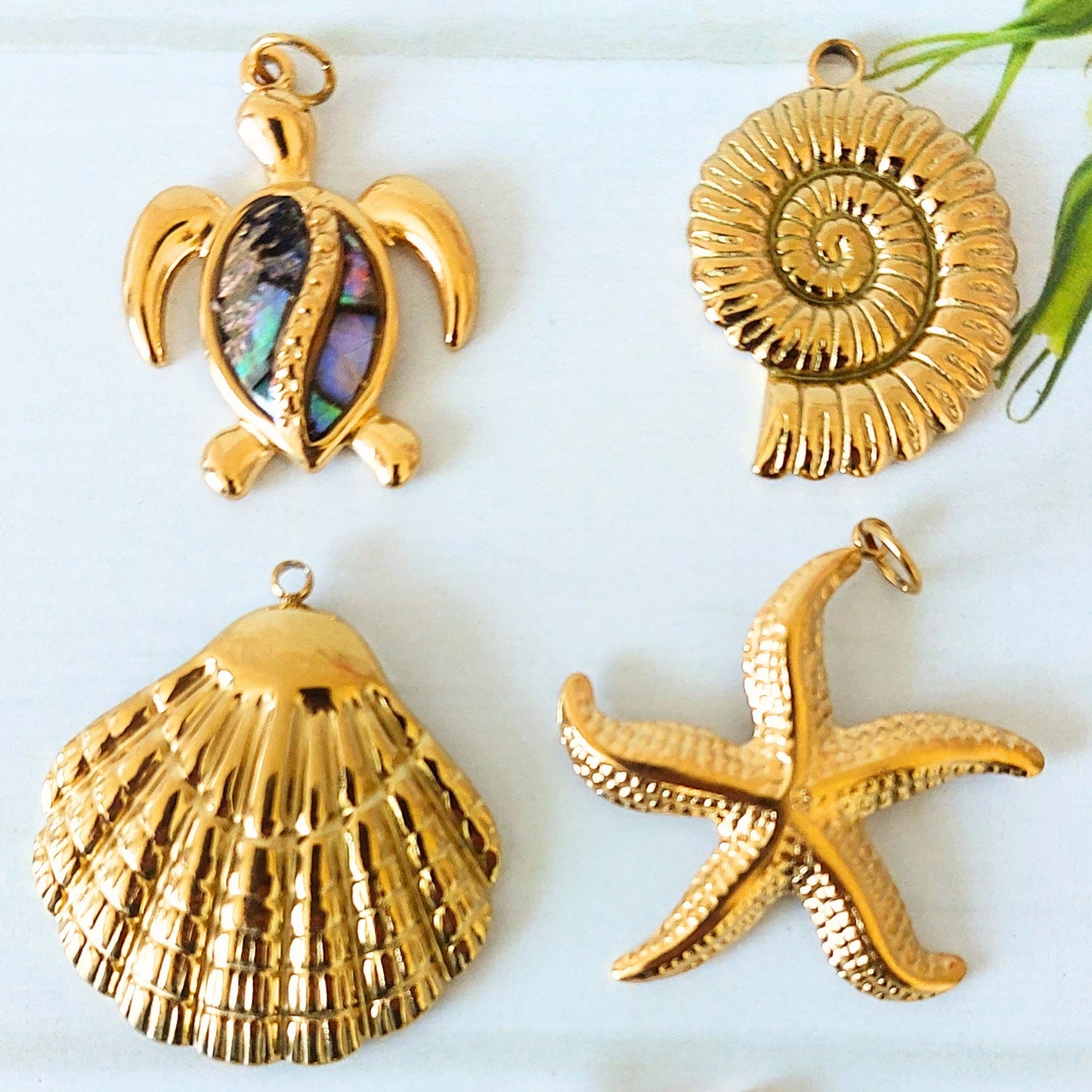 Four gold-plated stainless steel shell and sea life jewelry charms, including a turtle with an abalone shell inlay, a spiral ammonite shell, a scallop shell, and a starfish, arranged on a white background.