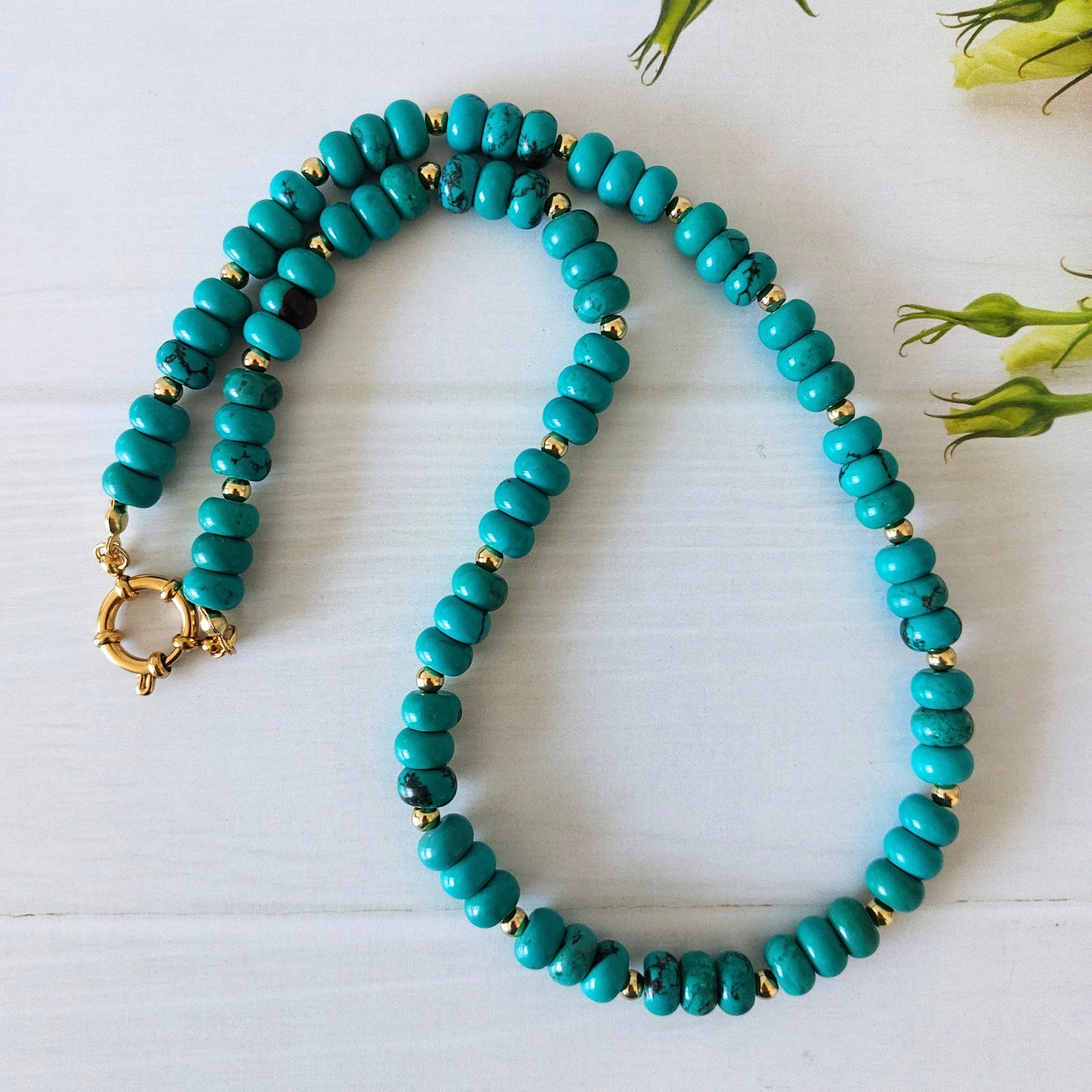 A turquoise coloured howlite rondelle bead necklace, accented by small gold-plated stainless steel spacer beads, displayed on a light wooden surface.