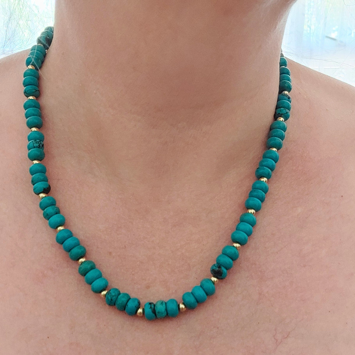 A turquoise coloured howlite beaded necklace with small gold-plated stainless steel spacer beads worn on a person's neck.