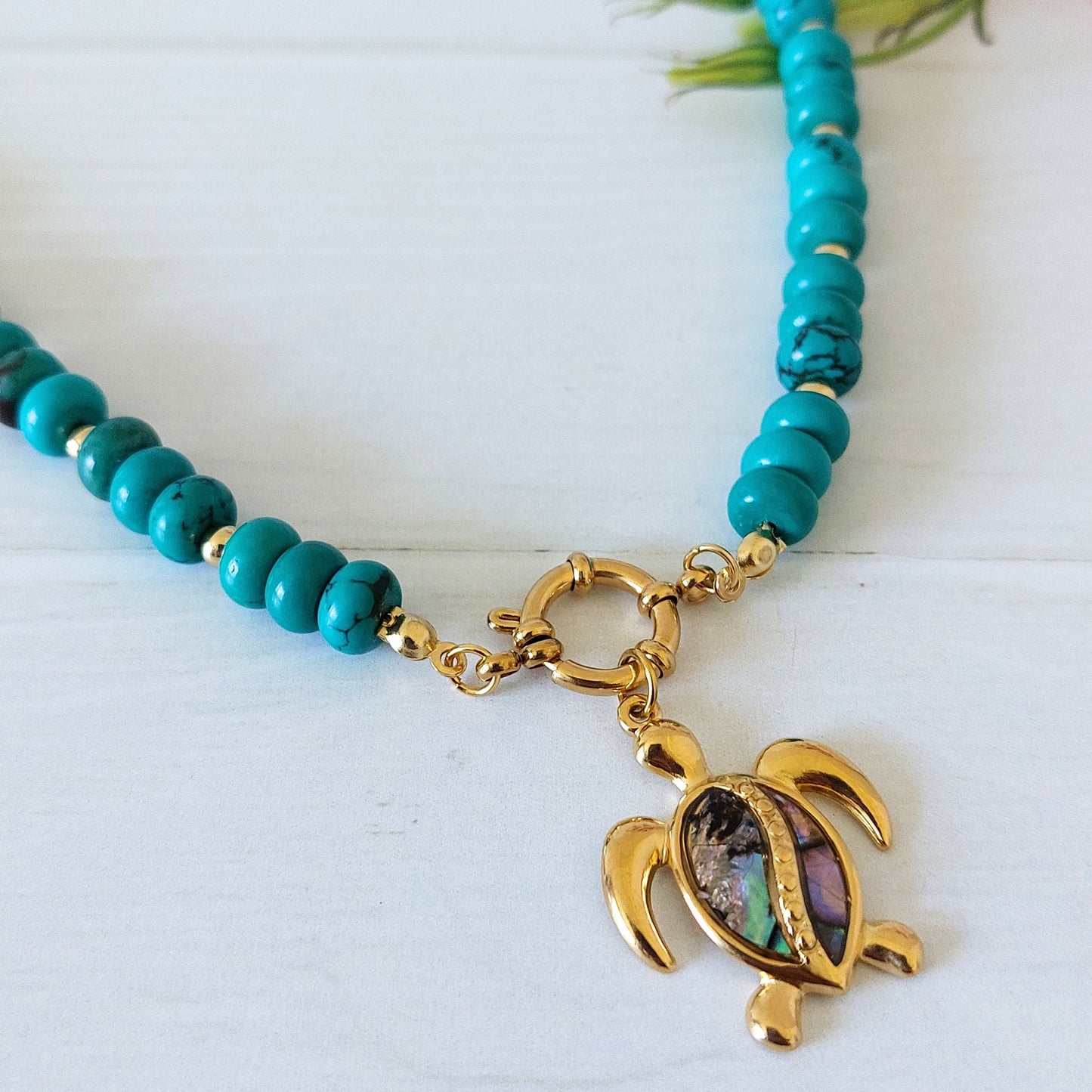 Close up of turquoise howlite bead necklace with a gold turtle pendant featuring an iridescent shell inlay, displayed on a white wooden surface.