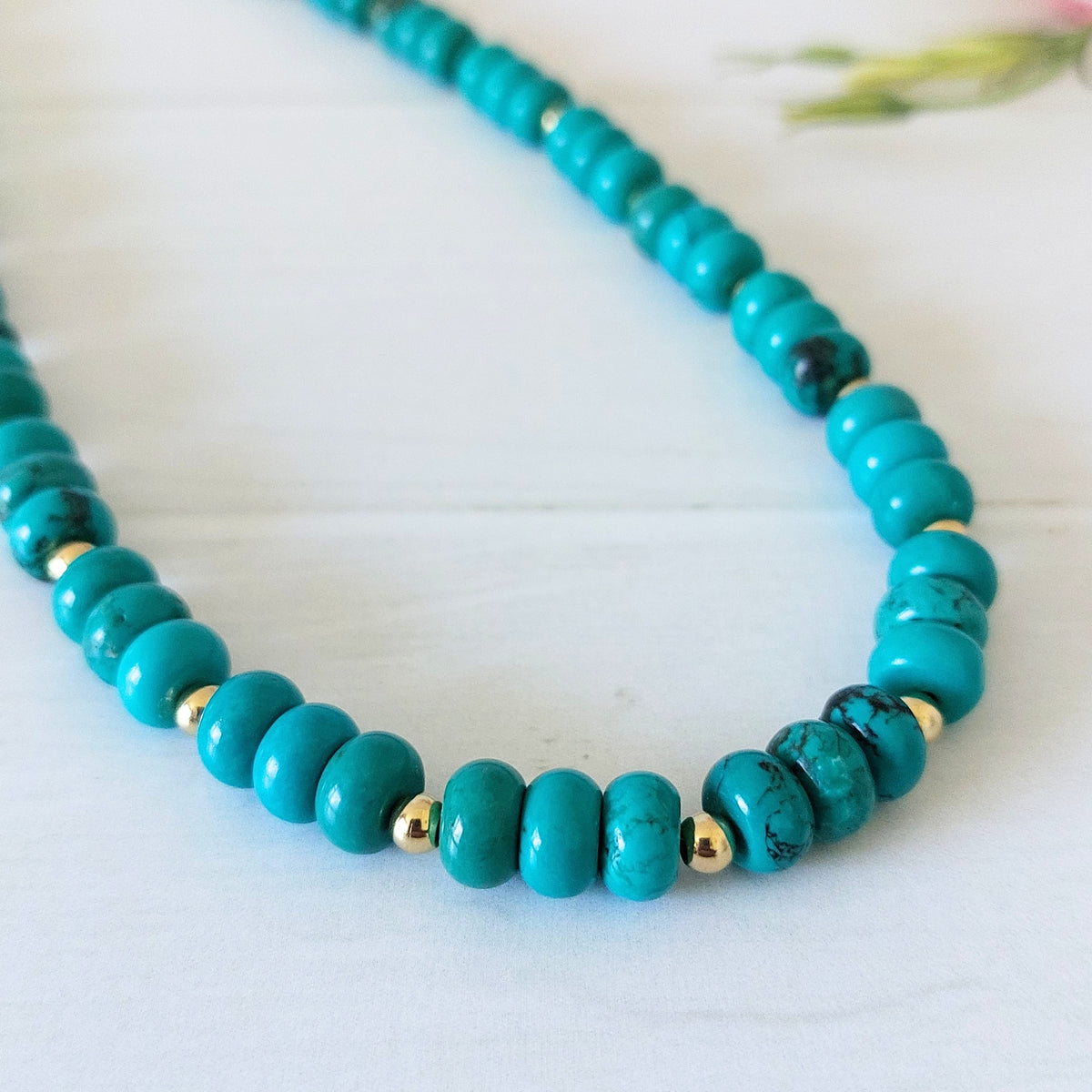 A turquoise coloured howlite rondelle bead necklace, accented by small gold-plated stainless steel spacer beads, displayed on a light wooden surface.