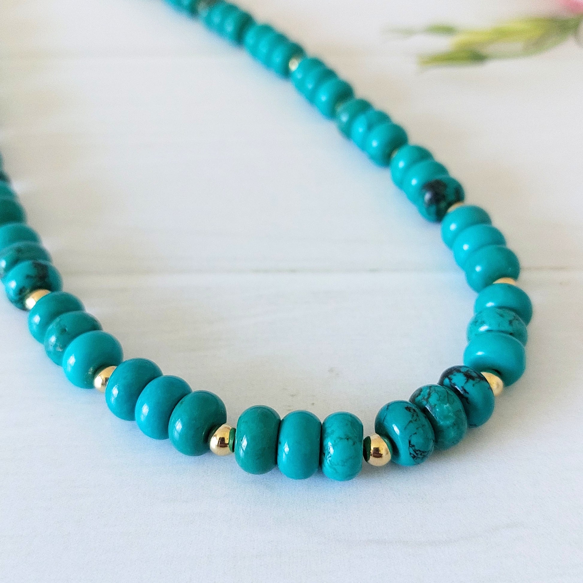 A turquoise coloured howlite rondelle bead necklace, accented by small gold-plated stainless steel spacer beads, displayed on a light wooden surface.