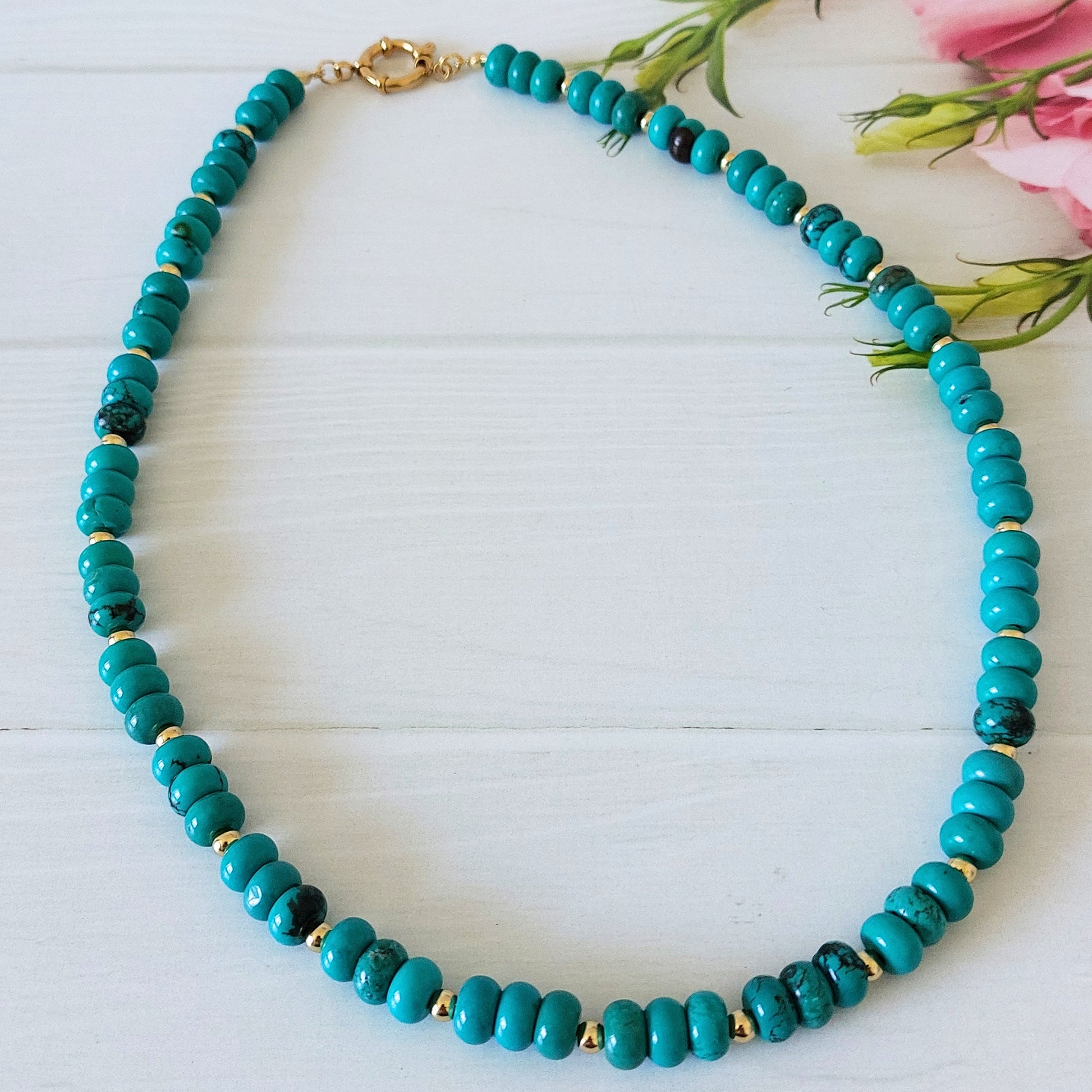 A turquoise coloured howlite rondelle bead necklace, accented by small gold-plated stainless steel spacer beads, displayed on a light wooden surface.