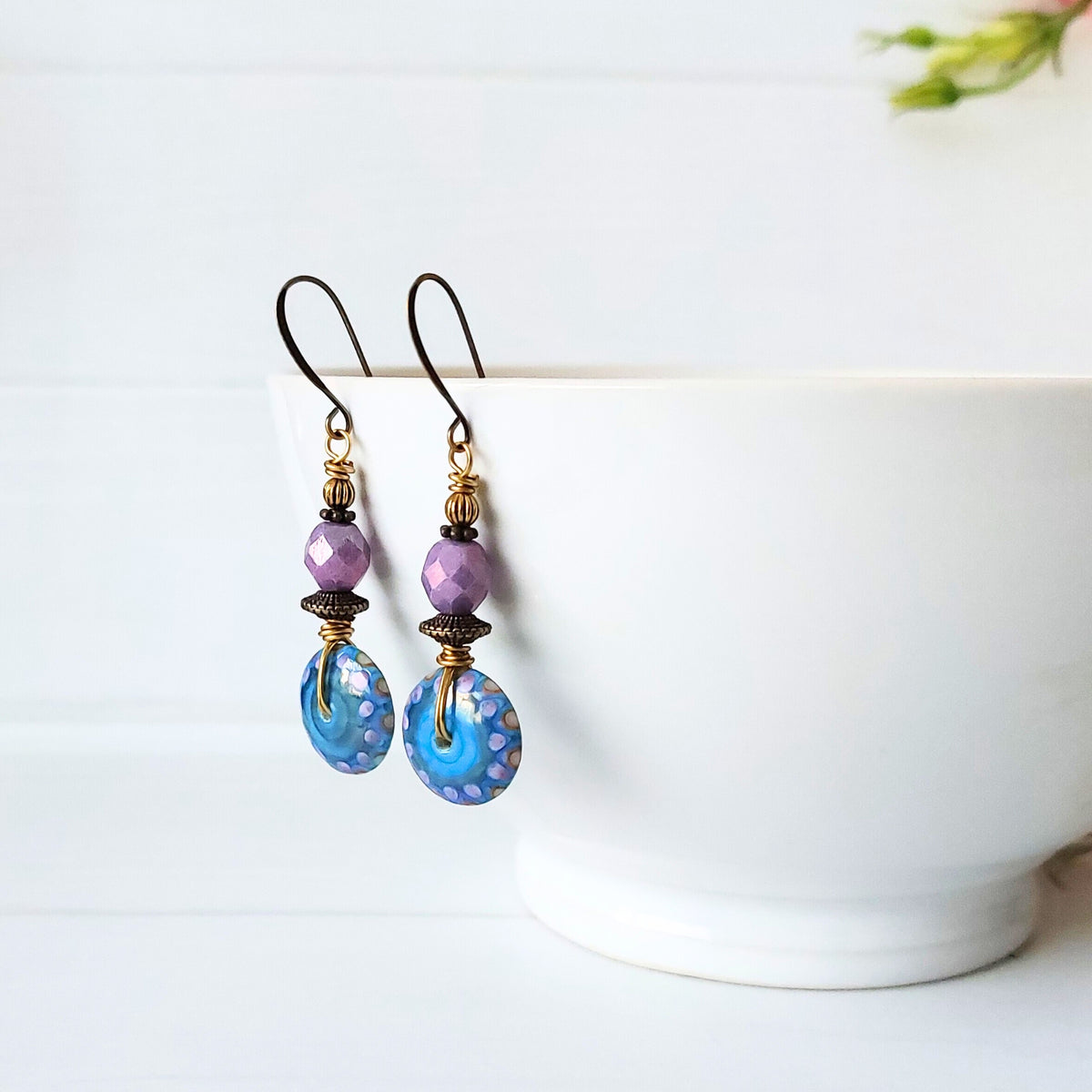 A pair of handmade dangle earrings with faceted purple beads, gold-toned spacers, and blue patterned glass disc charms, hanging in front of a white ceramic bowl on a white wooden surface.