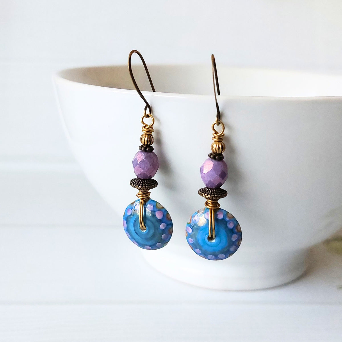 A pair of handmade dangle earrings with faceted purple beads, dark brass spacers, and large blue dotty glass disc beads, hanging in front of a white ceramic bowl.