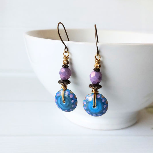 A pair of handmade dangle earrings with faceted purple beads, dark brass spacers, and large blue dotty glass disc beads, hanging in front of a white ceramic bowl.