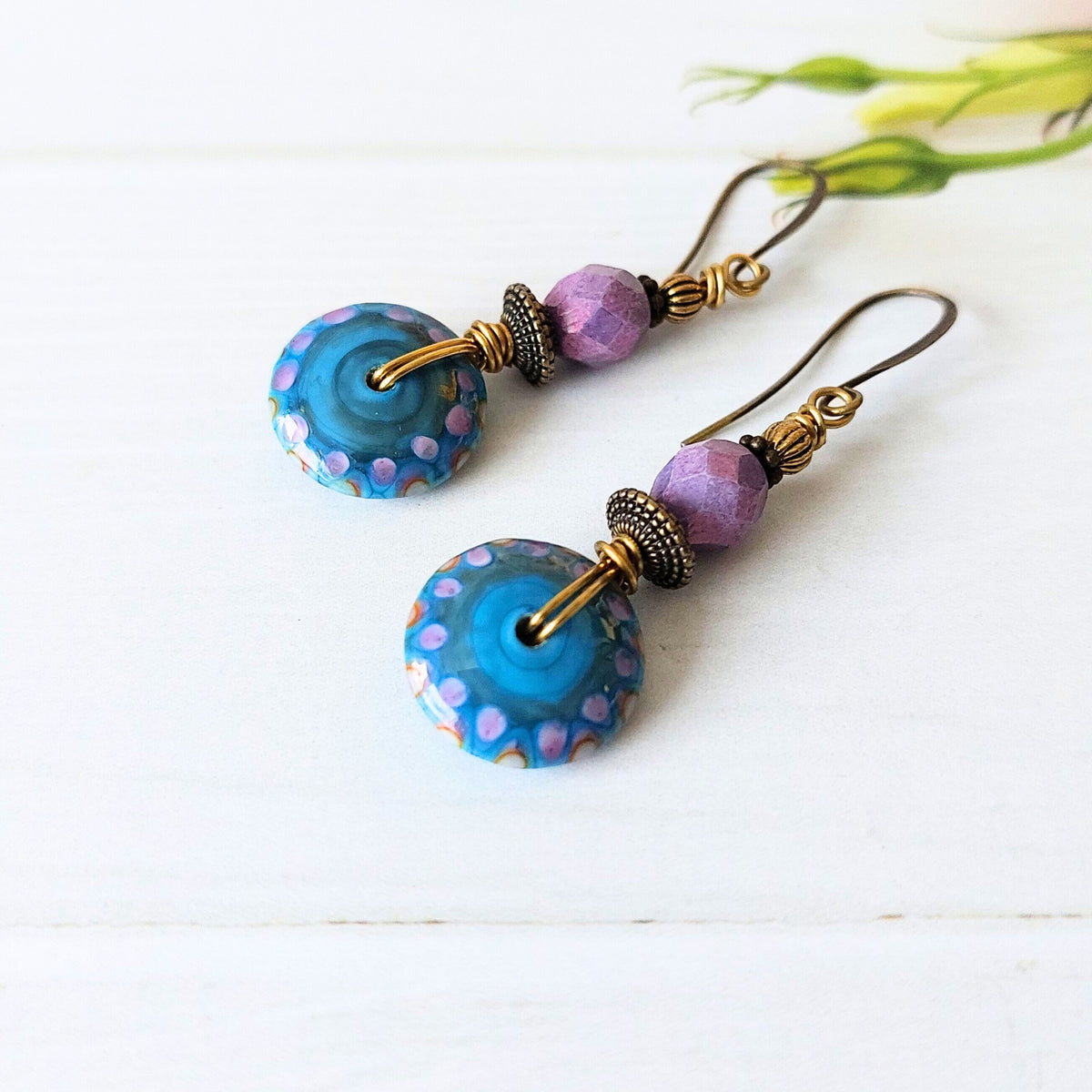 A pair of handmade dangle earrings featuring round blue and purple dots glass disc beads, faceted lavender beads, and gold-toned wire hooks, displayed on a white wooden surface.