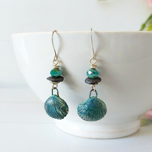 Handmade polymer clay shell drops hang from dark grey ceramic discs and ice-blue faceted glass beads on silver-plated earwires. Hanging from a white ceramic bowl.