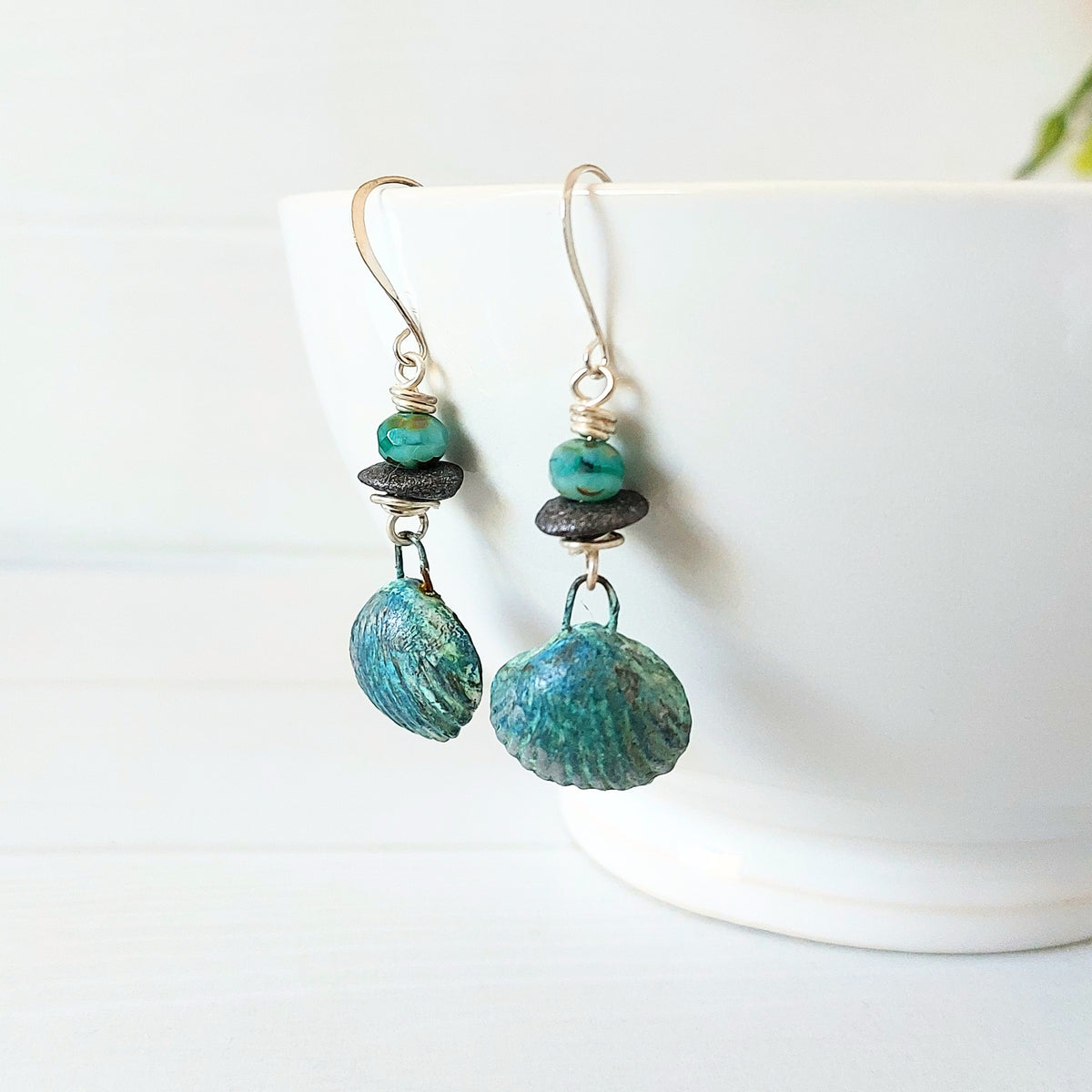 Handmade polymer clay shell drops hang from dark grey ceramic discs and ice-blue faceted glass beads on silver-plated earwires. Hanging from a white ceramic bowl.